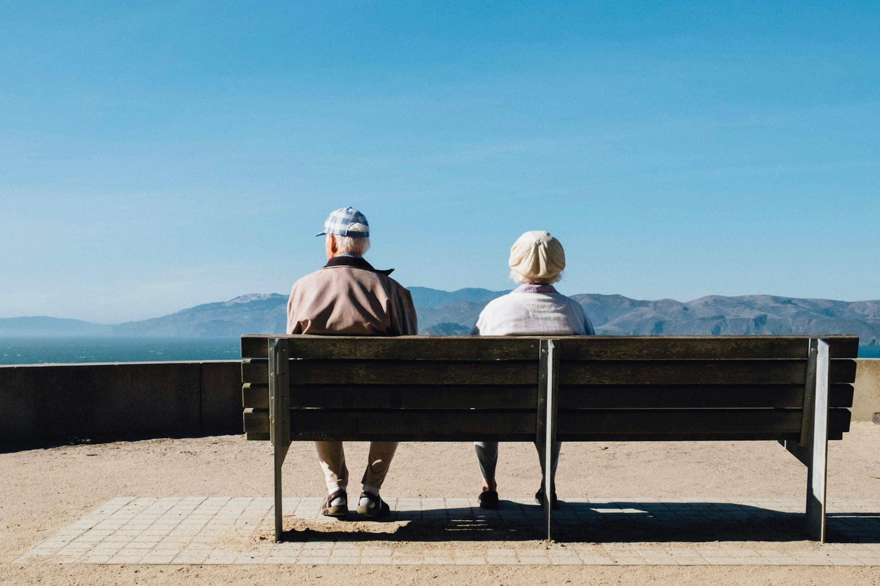An older couple sitting on a wooden bench, looking out over the water towards distant hills